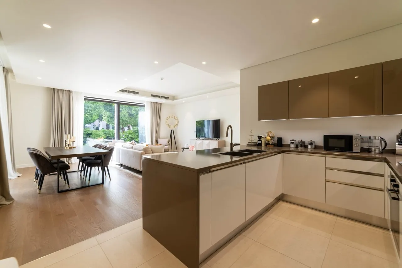 Modern kitchen with an island and brown cabinets; a dining area and sofa are visible in the background.
