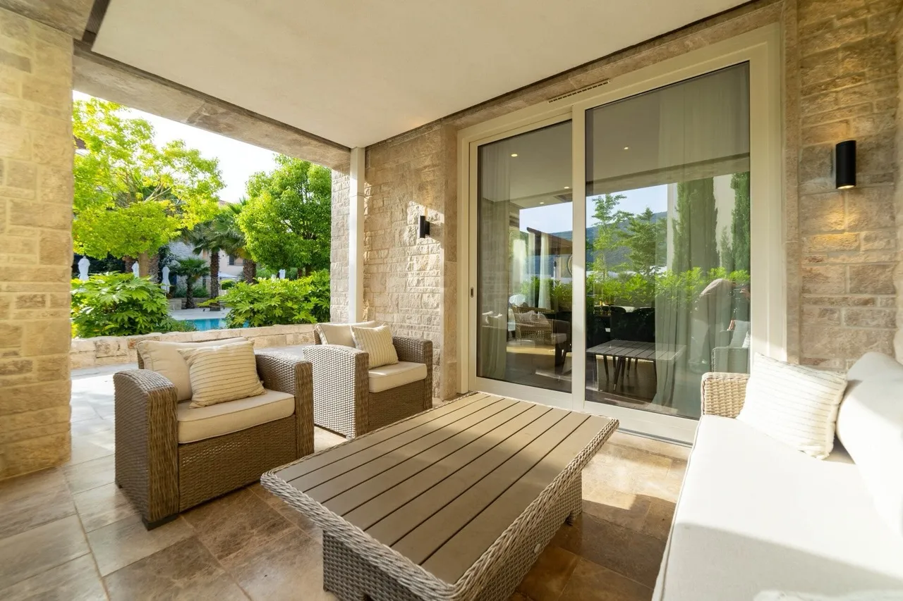 Covered terrace with wicker furniture, stone walls, sliding glass doors, and garden view.