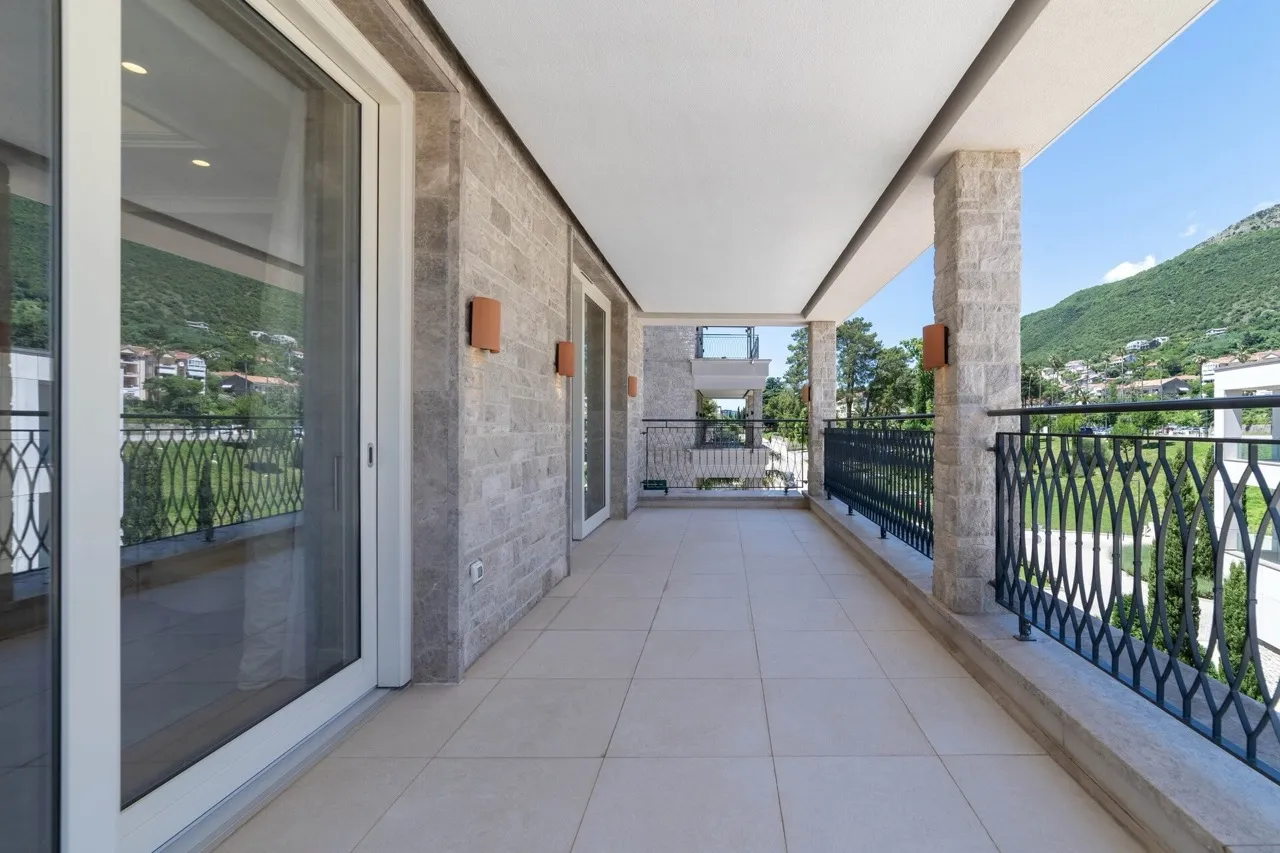 Long balcony with stone wall, black metal railing, and view of green hills under a covered roof.