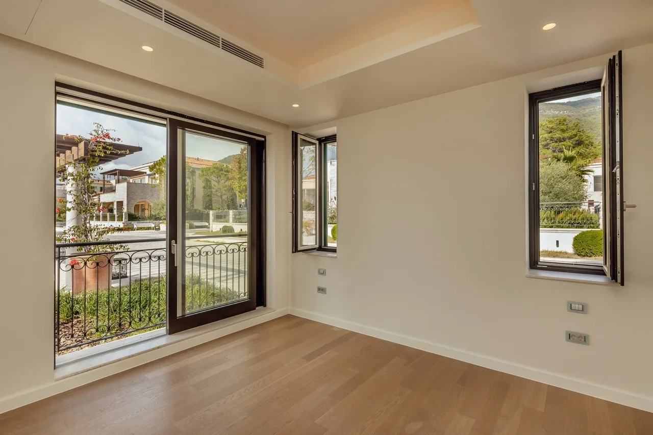 Empty room with wooden flooring and large windows opening to a balcony overlooking a garden and neighboring houses.