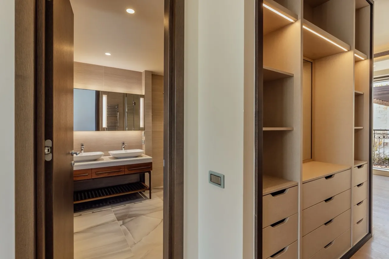 Walk-in closet with built-in shelving and drawers; bathroom visible through doorway with double sinks.