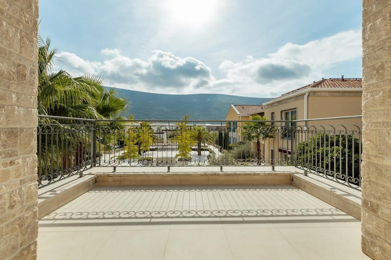 Stone balcony with wrought iron railing overlooking palm trees, neighboring houses, and mountains under a cloudy sky.