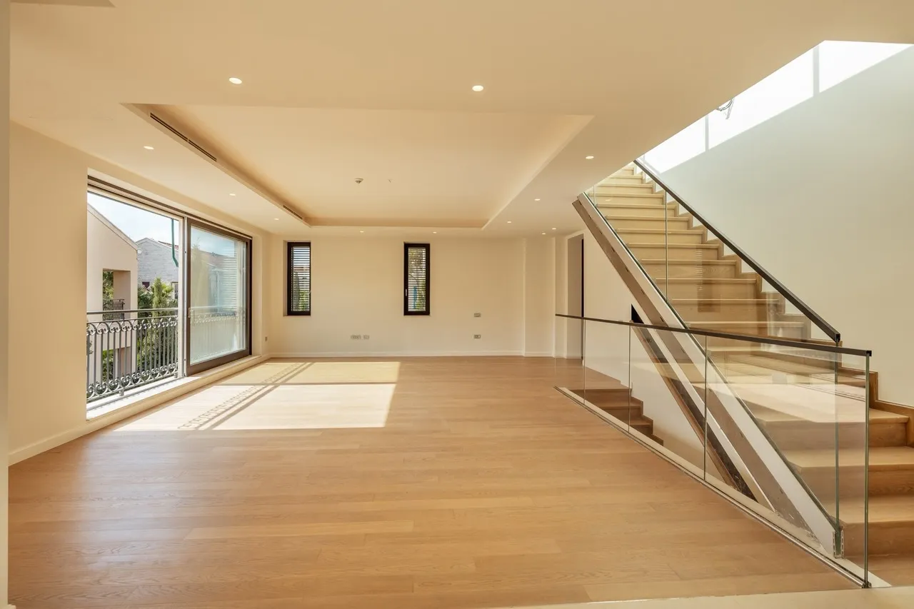 Empty room with wooden flooring, a staircase with glass railings, and a large window leading to a balcony.