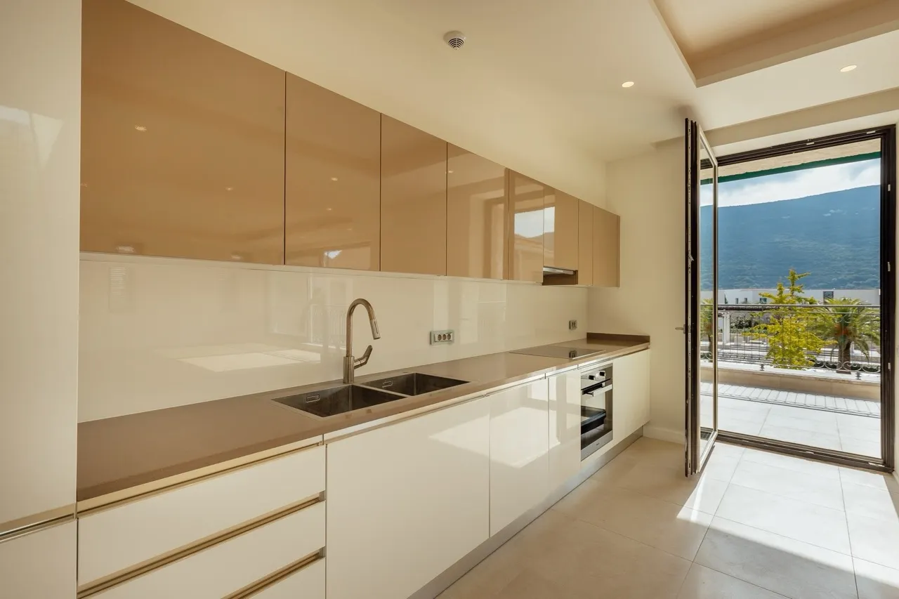 Modern kitchen featuring beige glossy cabinets, a sink, cooktop, and an open door leading to a balcony with mountain views.