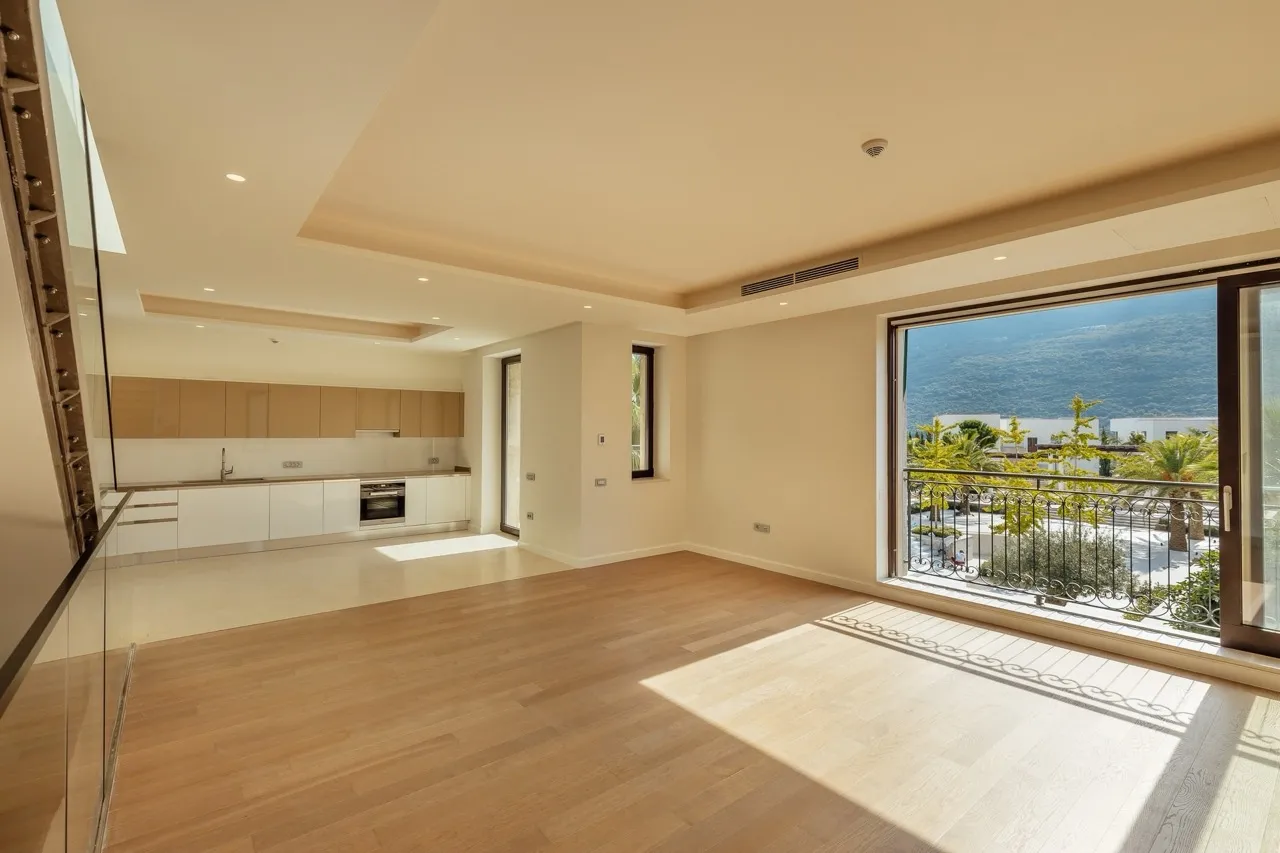 Bright open room with wood flooring, visible kitchen in background, and sliding door leading to a balcony with mountain views.