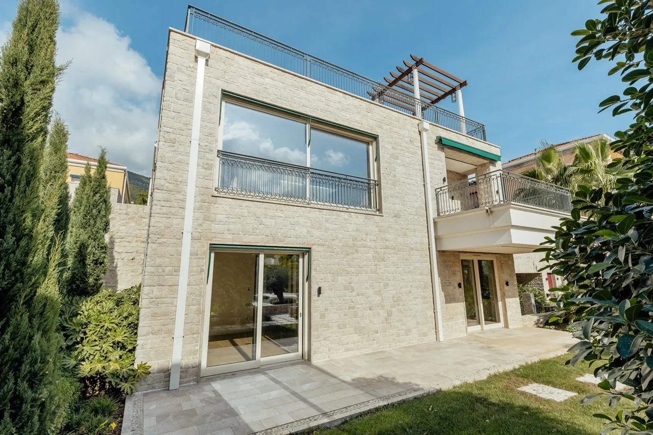 Exterior view of a two-story house with light stone masonry, balconies, and glass doors under a blue sky.