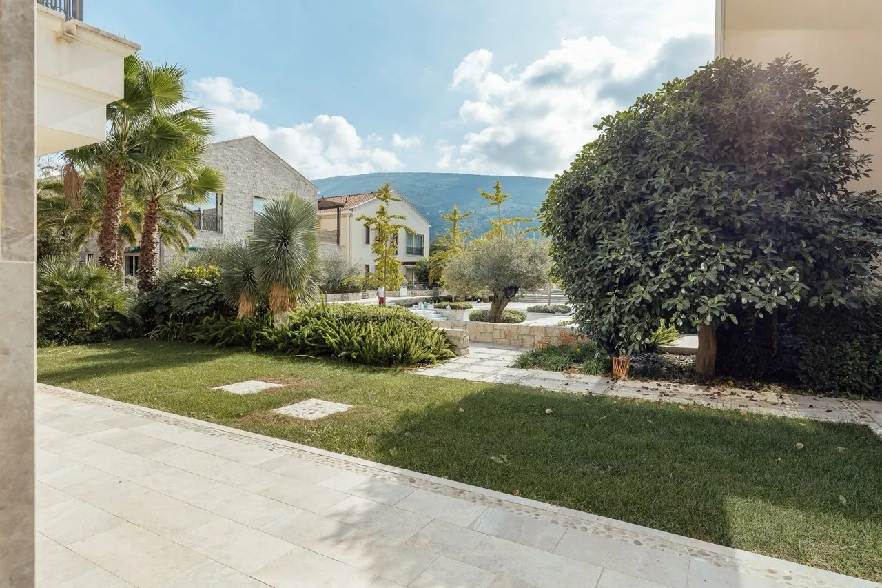 View of a garden with lawn, palm trees, and stone path. Other houses and mountains are visible in the background under a cloudy sky.