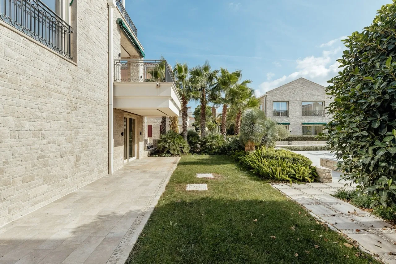 Stone house facade with balcony, garden path, green lawn, and palm trees under a clear blue sky.