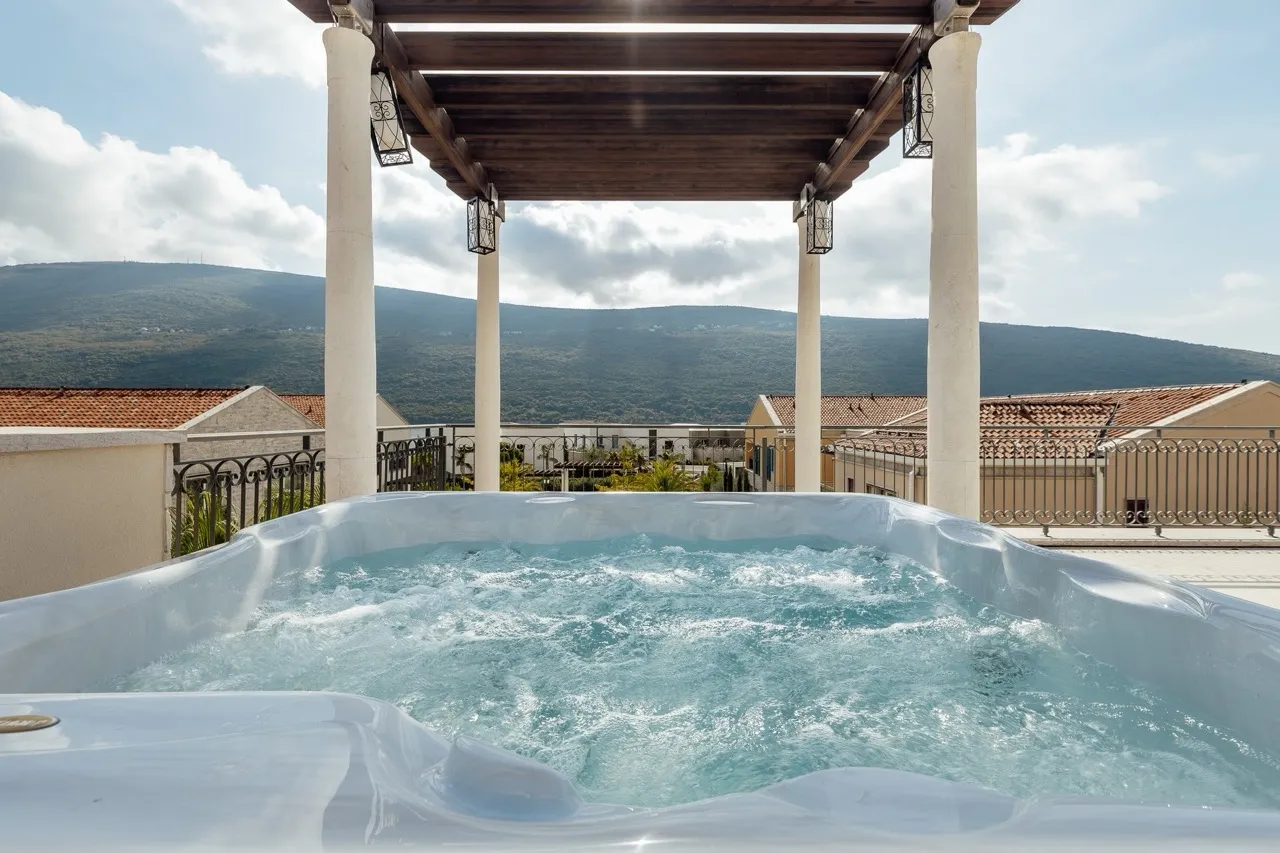 White bubbling jacuzzi on a terrace under a wooden pergola with a mountain view in the background.