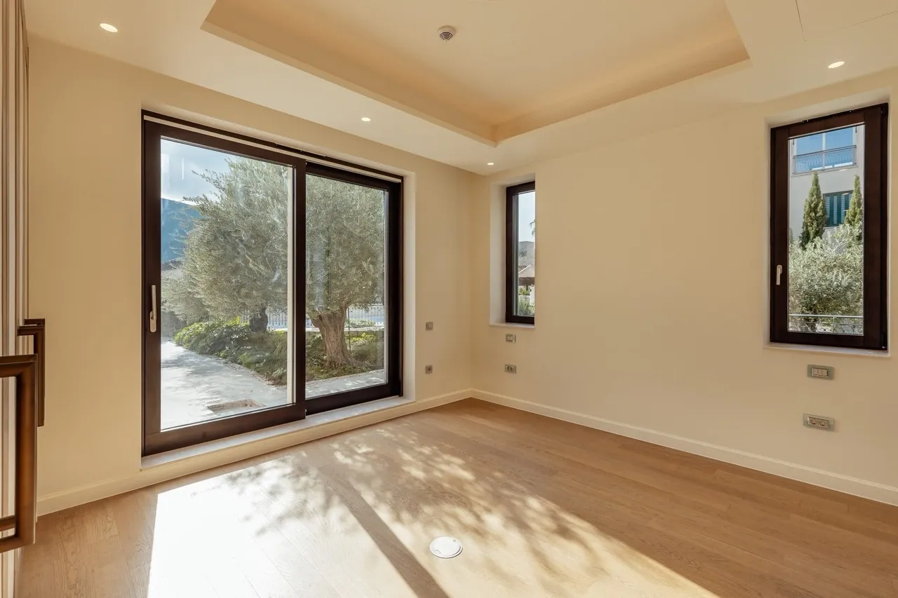 Empty room with wooden flooring, large windows overlooking a garden. Light walls and recessed ceiling lights.