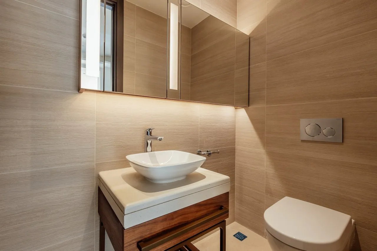 Modern bathroom featuring a vessel sink on a wooden vanity, wall-mounted toilet, and beige tiled walls with mirror lighting.