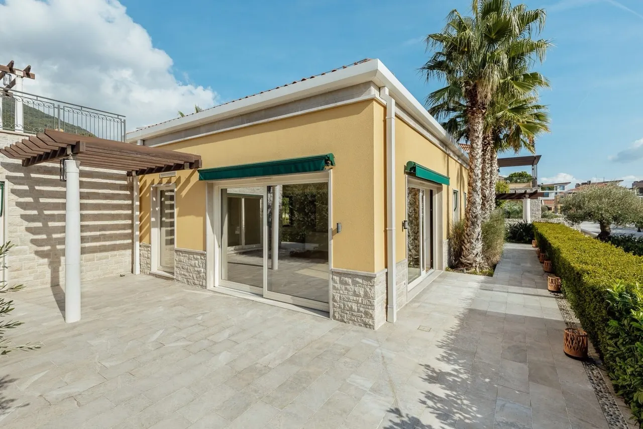 Yellow house facade with stone patio, wooden pergola, and palm tree under a blue sky.