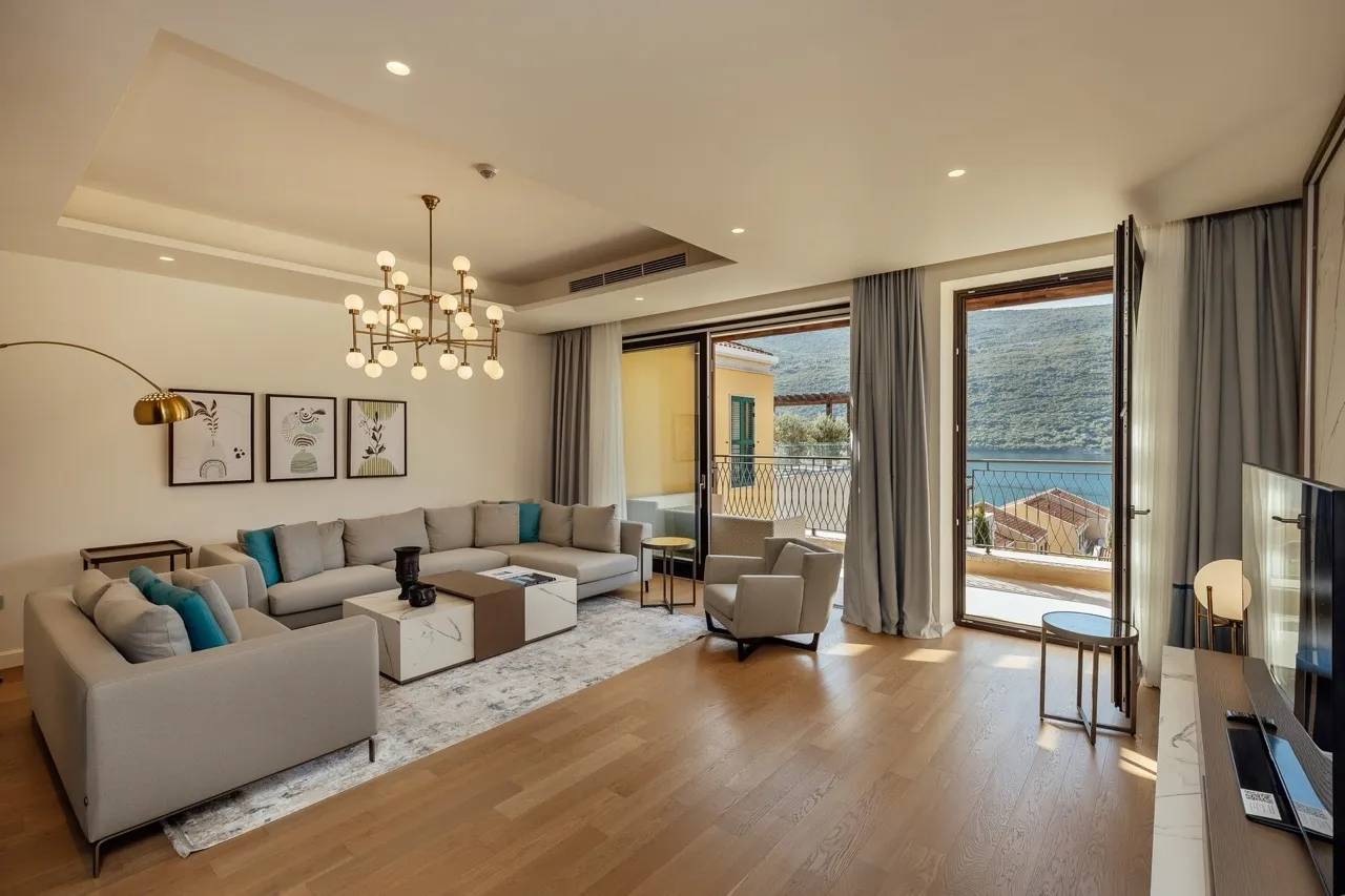 Bright living room with a grey sectional sofa, wooden floors, and open doors leading to a balcony overlooking water.