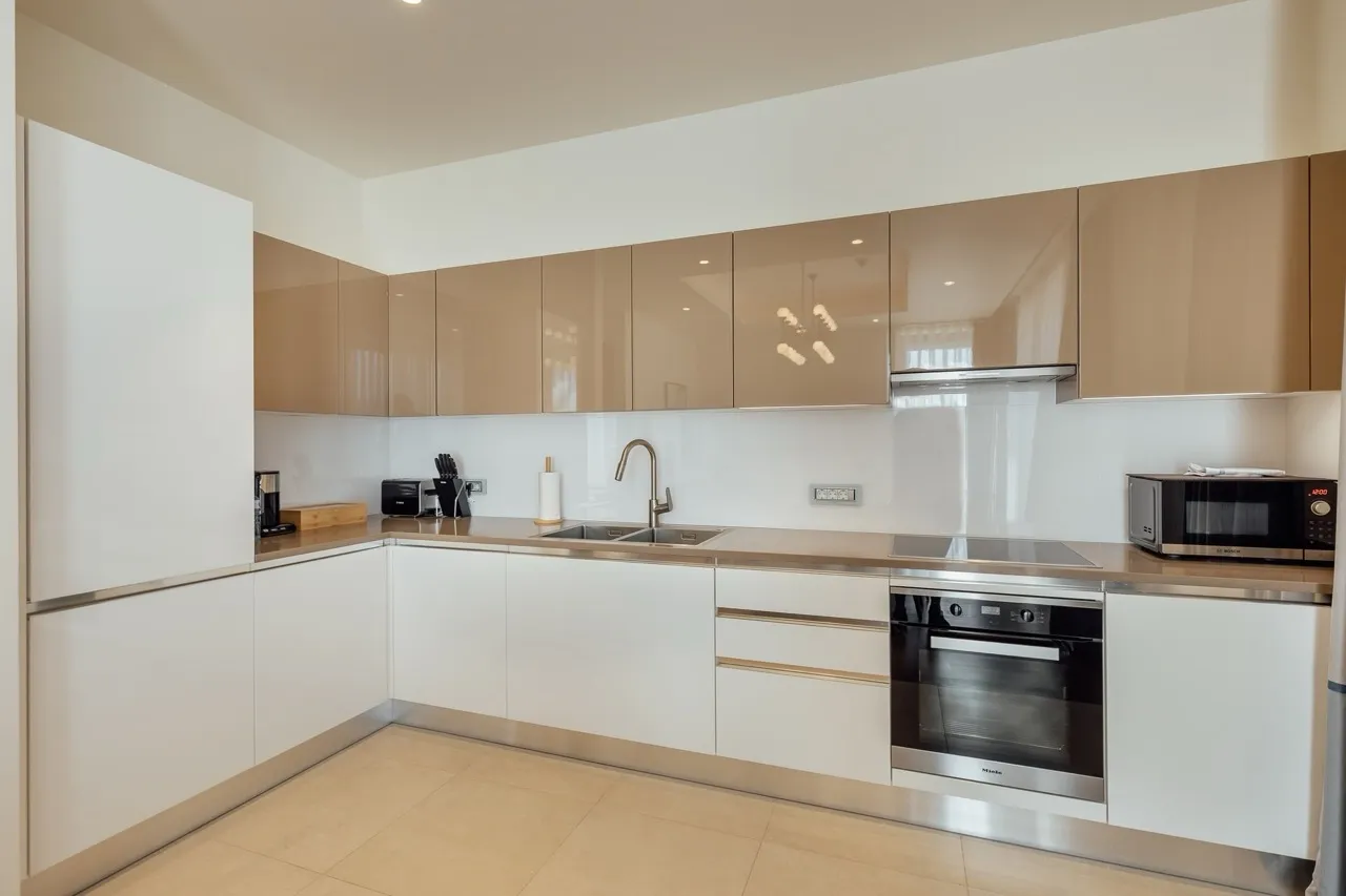 Modern kitchen with white and beige glossy cabinets, built-in Miele oven, sink, microwave, and stainless steel countertops.
