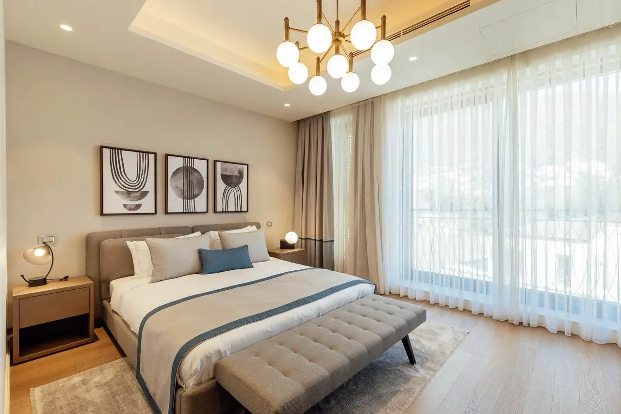 Modern bedroom featuring a double bed, nightstands, a tufted bench, abstract wall art, and large windows with sheer curtains.