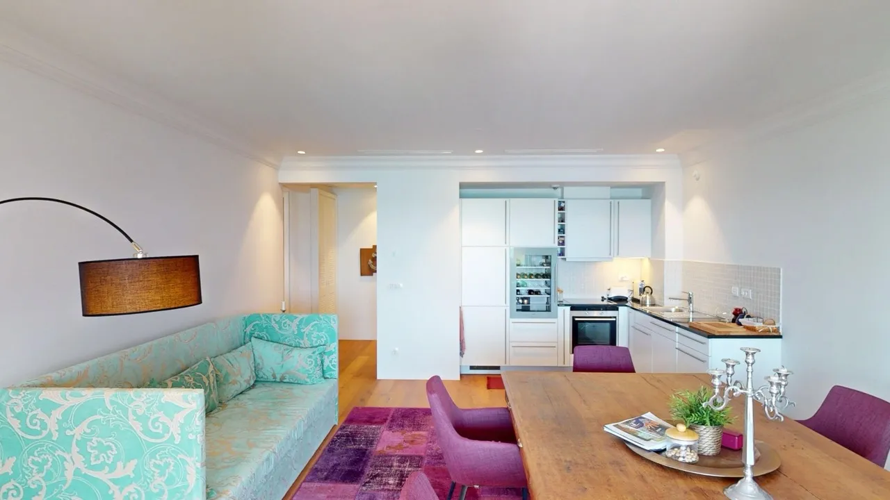 Living room with a turquoise sofa and wooden dining table, open kitchen visible in background.