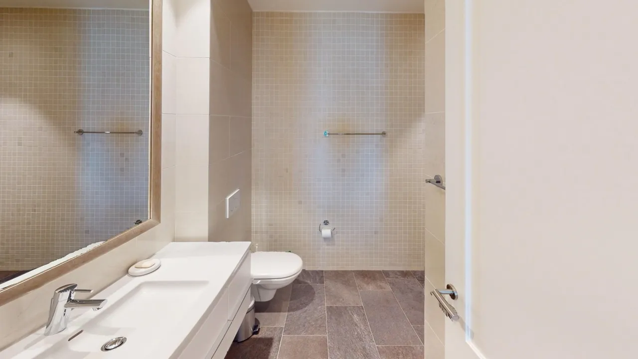 Bathroom featuring a white vanity sink, wall-mounted toilet, large wooden-framed mirror, and beige mosaic wall tiles.