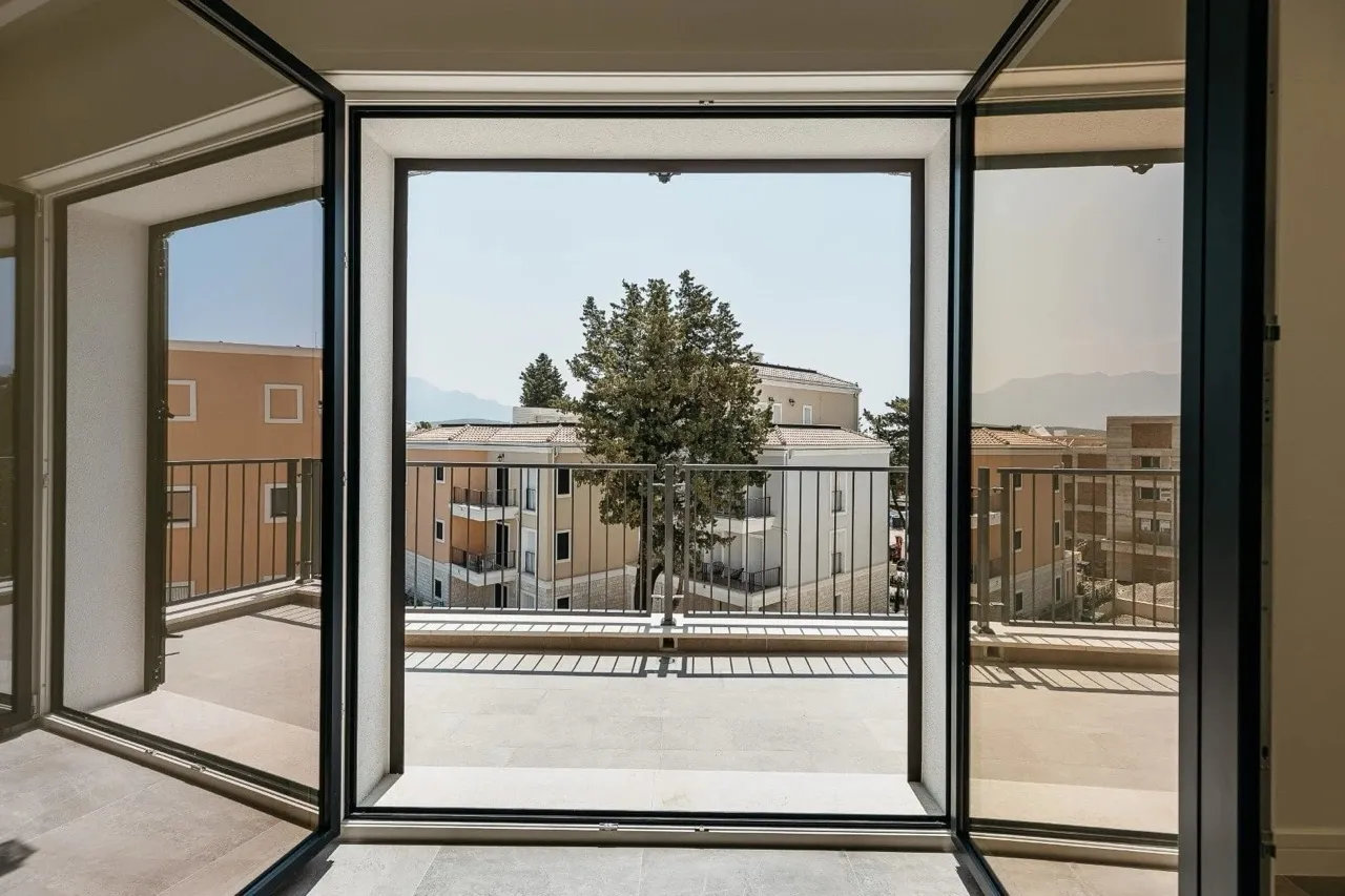 Open black doors lead to a concrete balcony with a metal railing overlooking nearby buildings.