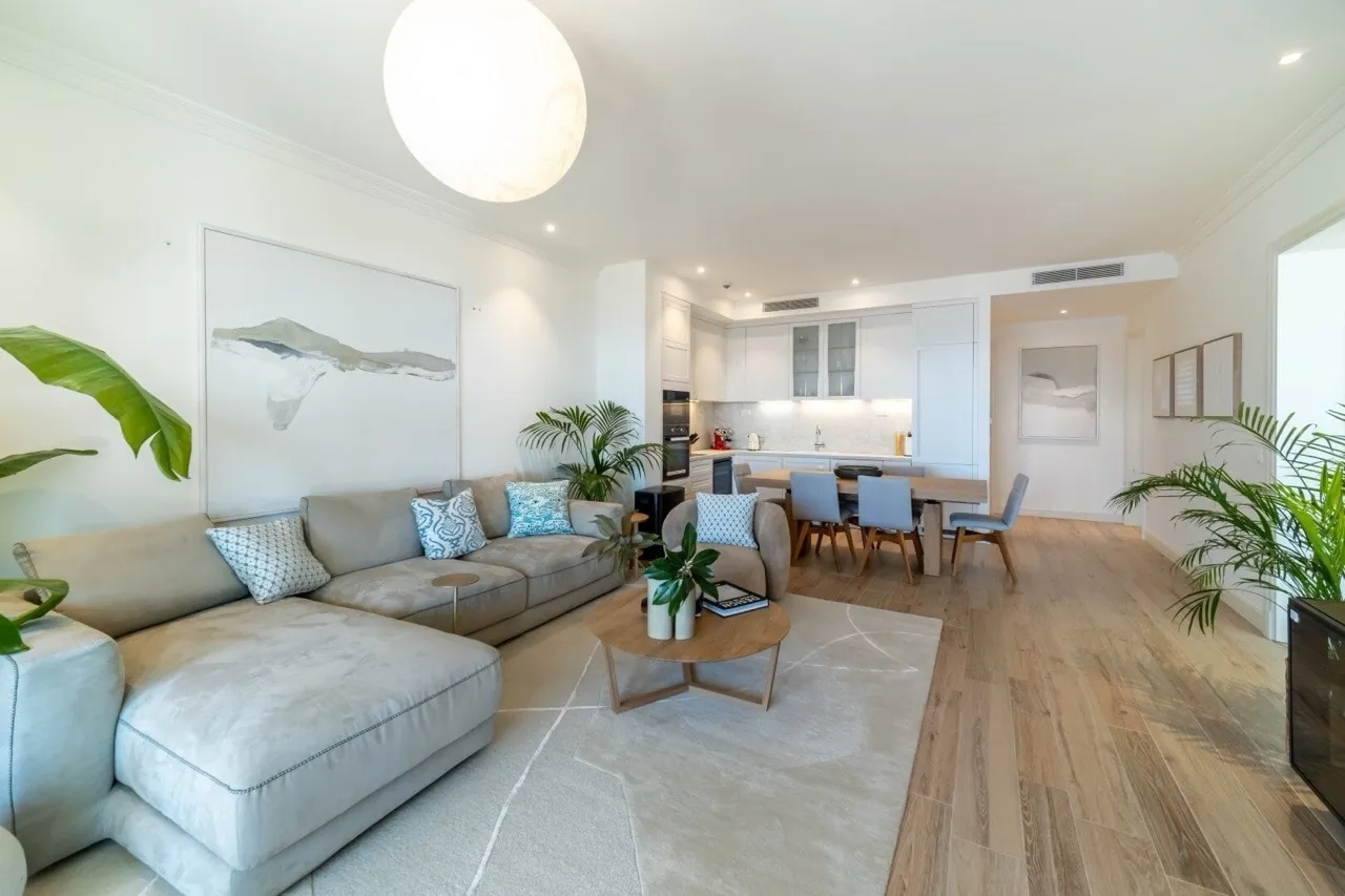 Bright living room with a grey sectional sofa, wooden dining table, and visible kitchen area in the background.