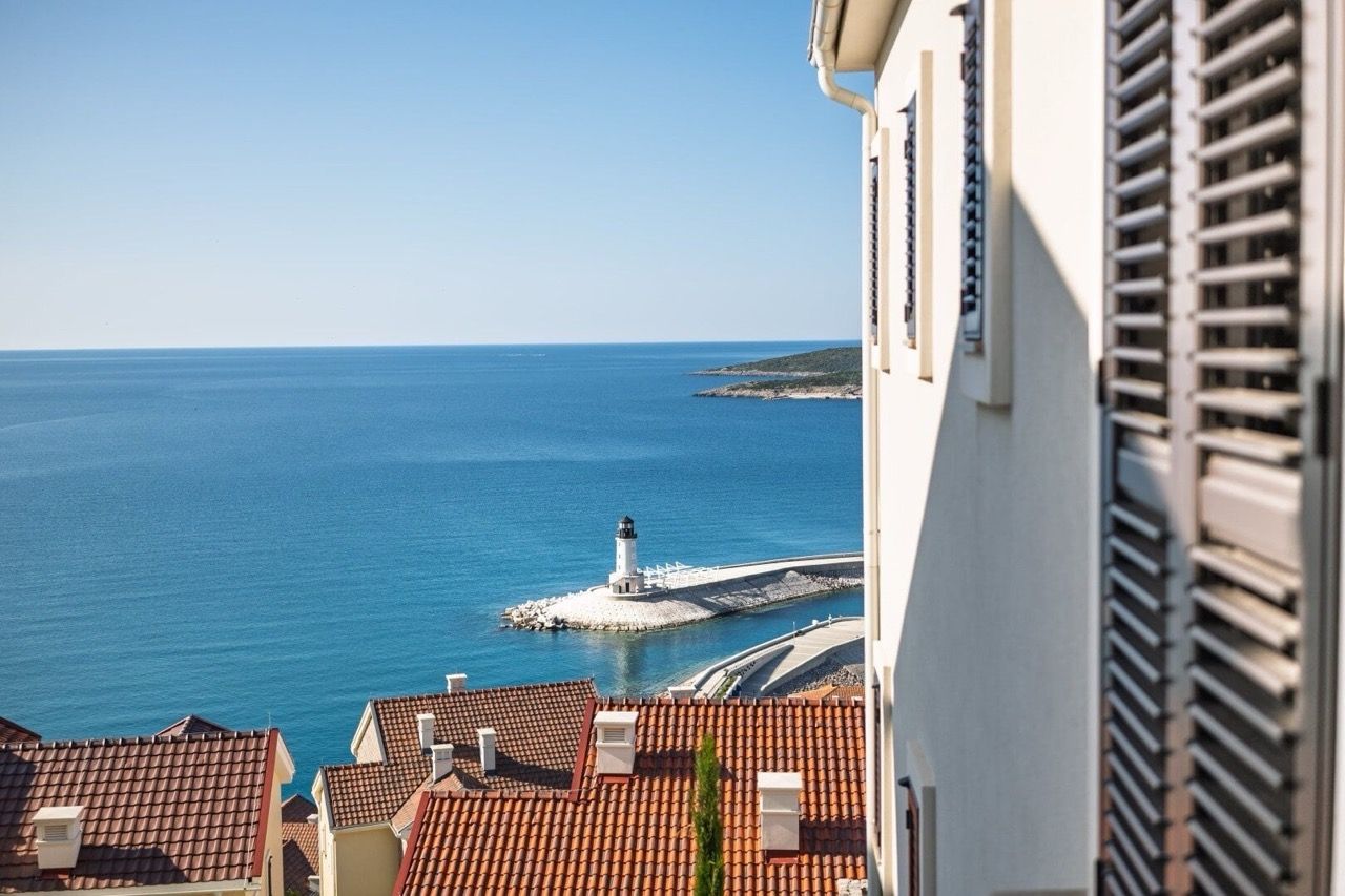 View from the apartment window in Luštica Bay, Radovići, Montenegro: the sea, a lighthouse, and the tiled roofs of houses on the coast.