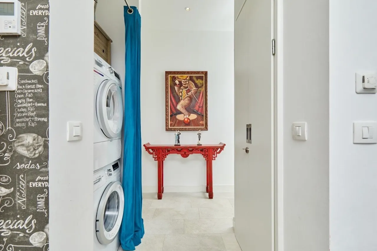 Washing machine behind blue curtain, red console table with painting in a hallway.