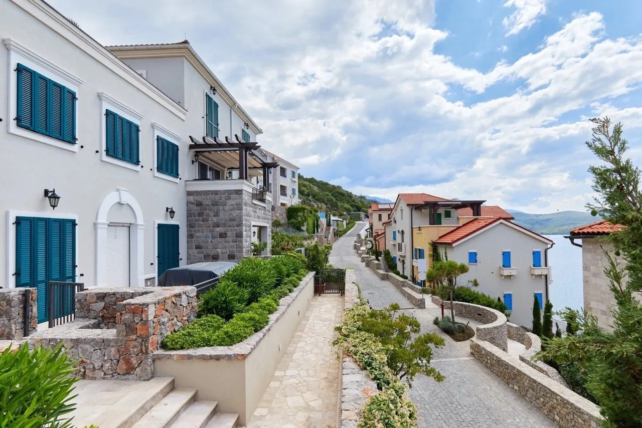 Stone pathway between white houses with blue shutters and greenery near the water.