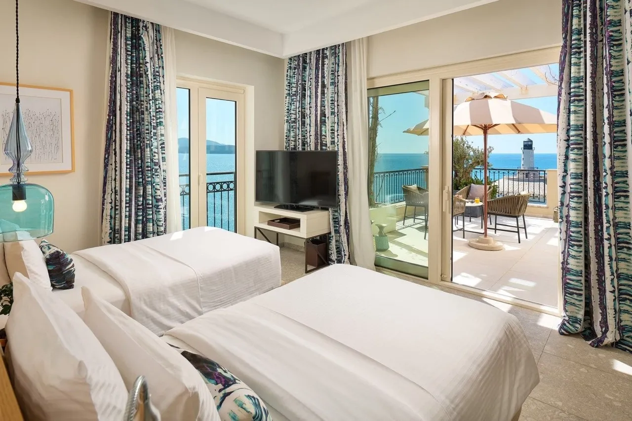 Two white beds, TV, and sliding doors opening to a balcony with sea and lighthouse view.
