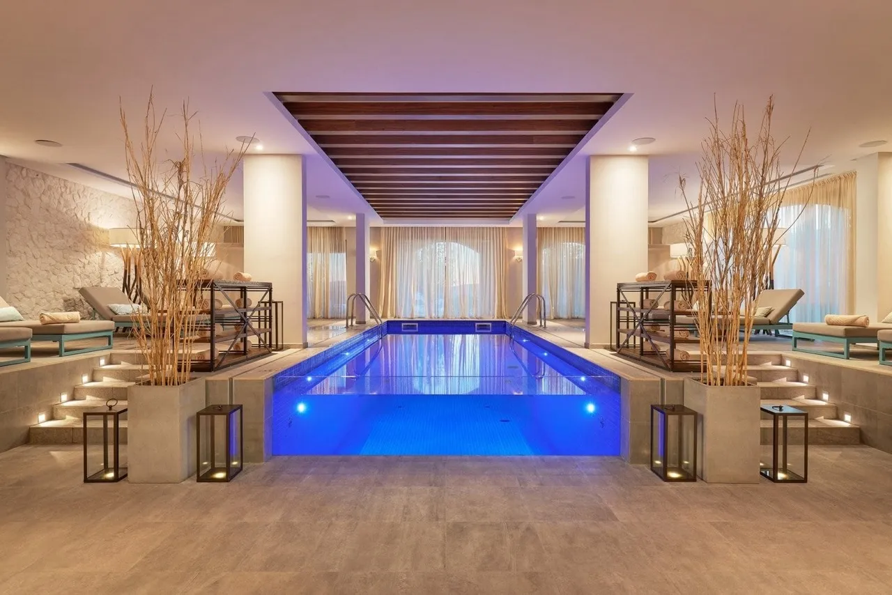 Bright indoor pool with blue underwater lighting, surrounded by lounge chairs and decorative dried reed arrangements.