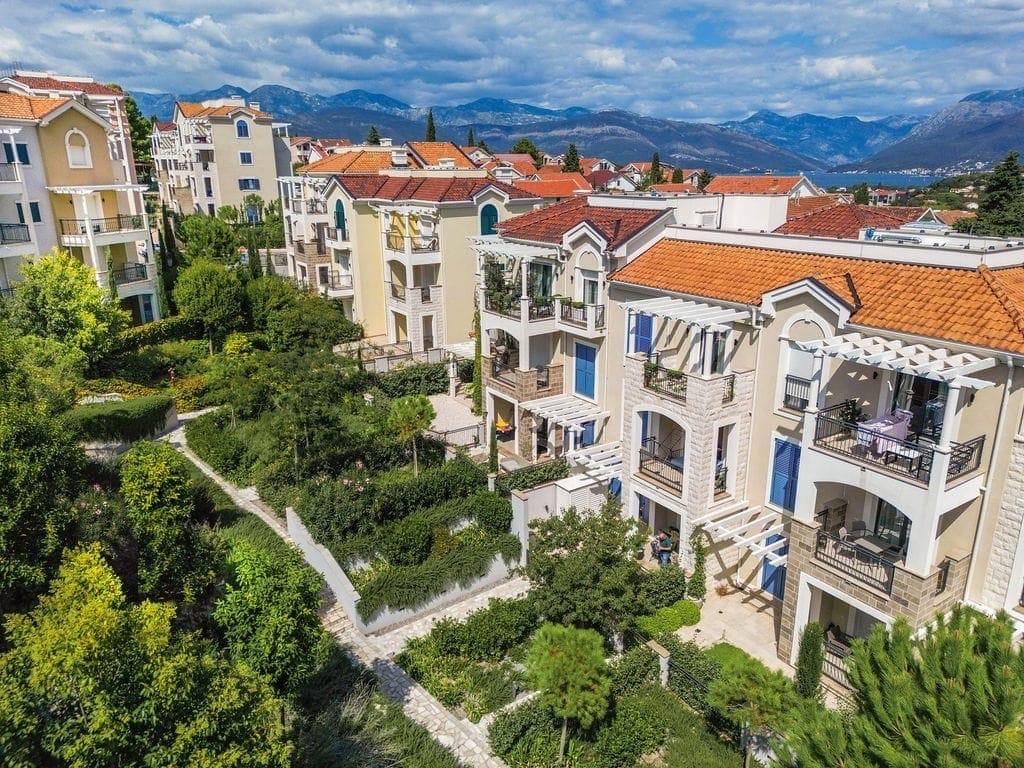 Aerial view of residential buildings with terracotta roofs, gardens, and mountains in the background.