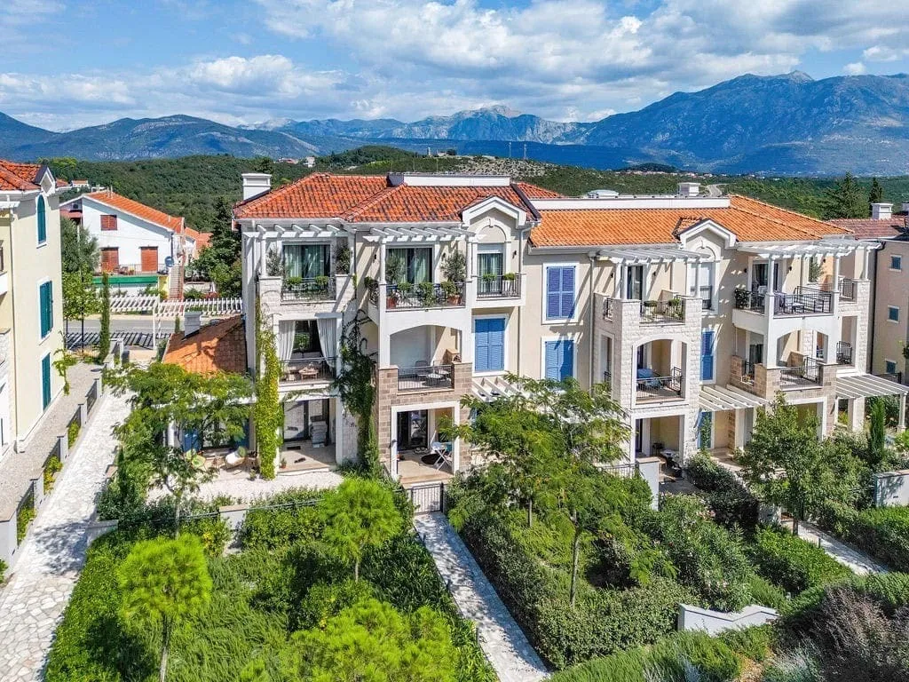 Aerial view of a multi-story house with terracotta roof, balconies, garden, and mountains.