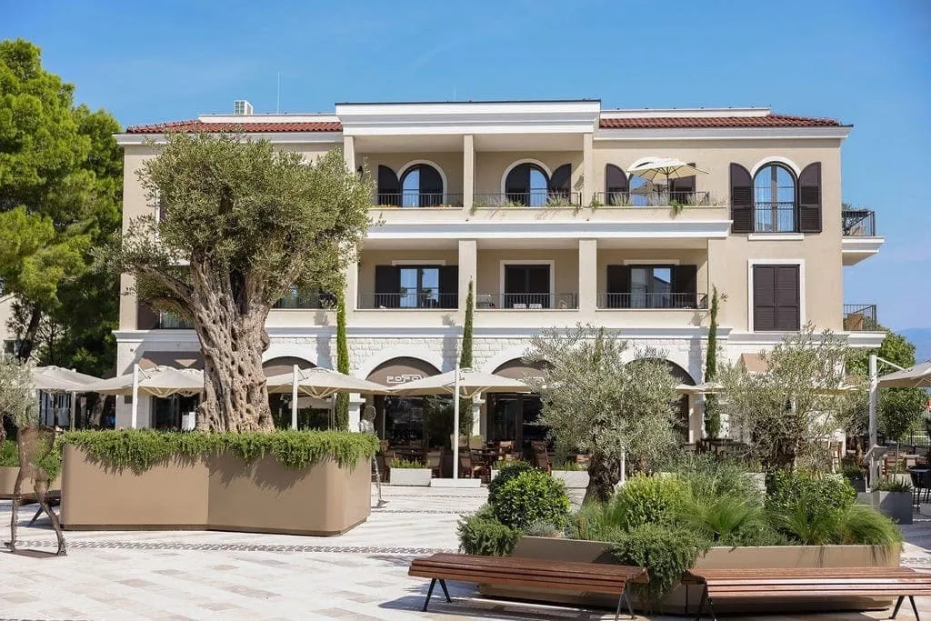 Exterior view of a multi-story building with a terrace, umbrellas, and an old tree in the foreground.