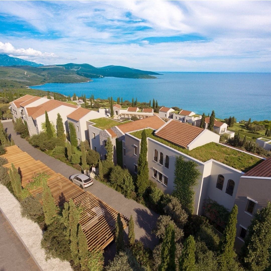 Aerial view of a building complex with terracotta roofs near the sea, surrounded by greenery and mountains.