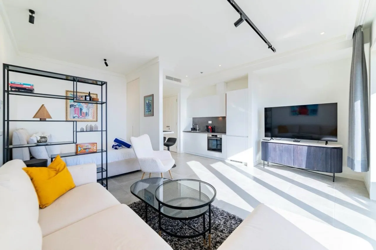 Bright studio with white sofa, TV, glass table, and open kitchen in background.