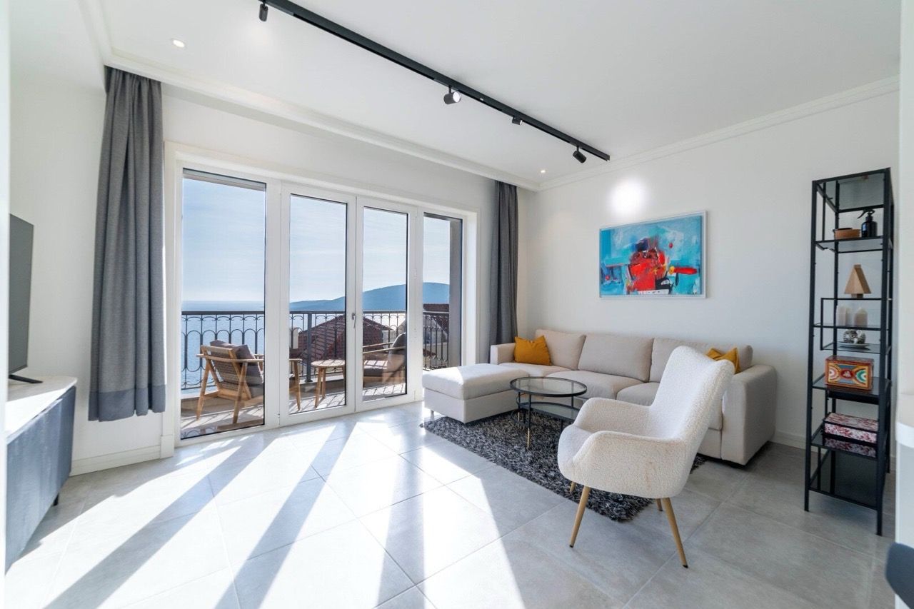 Bright living room with a sofa, armchair, and glass doors leading to a balcony overlooking the sea.