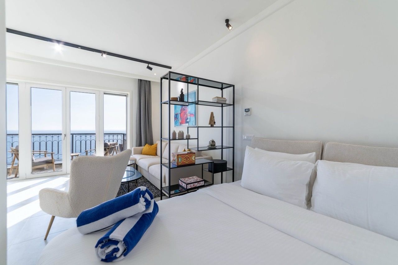 Bedroom featuring a large bed, white sofa, and metal shelving unit. A balcony door overlooks the sea.