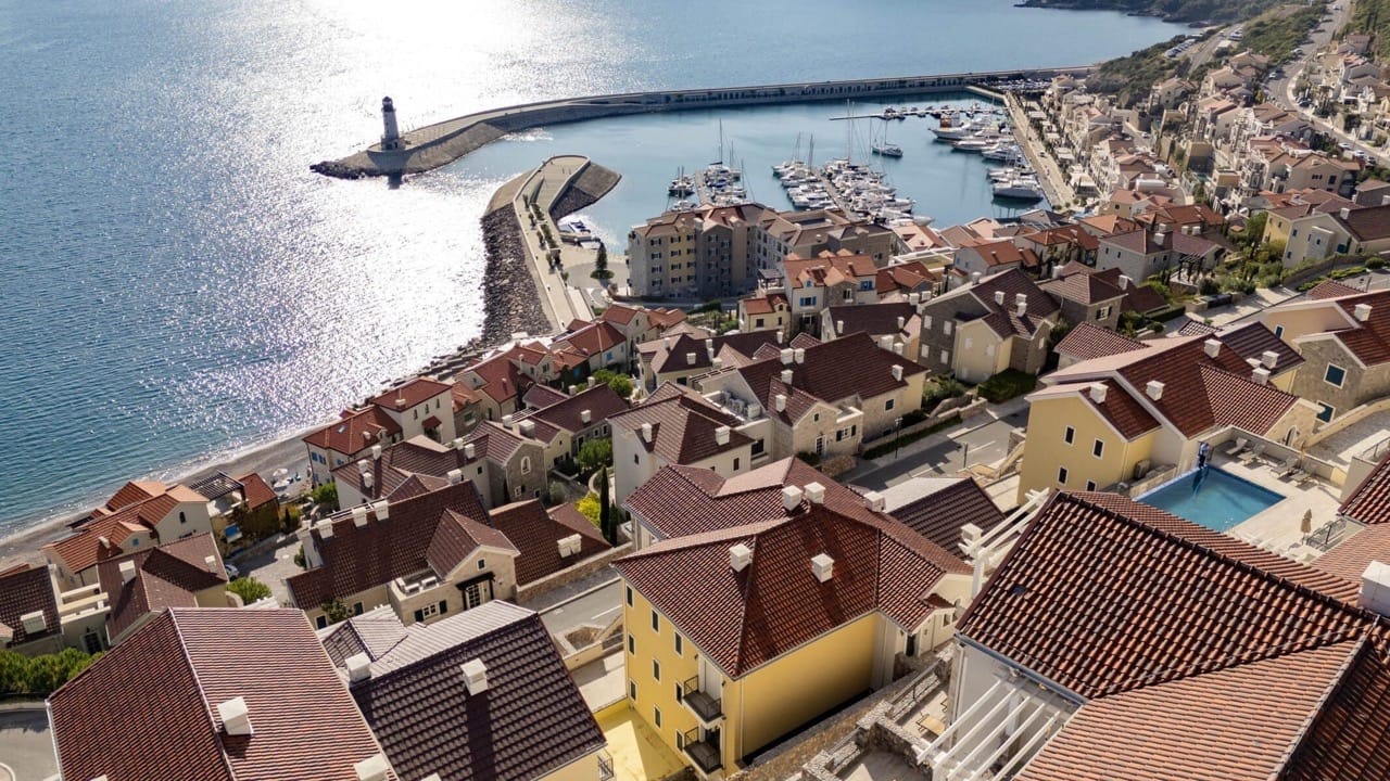 Aerial view of a coastal town showing red rooftops, a marina with boats, and a lighthouse by the sea.