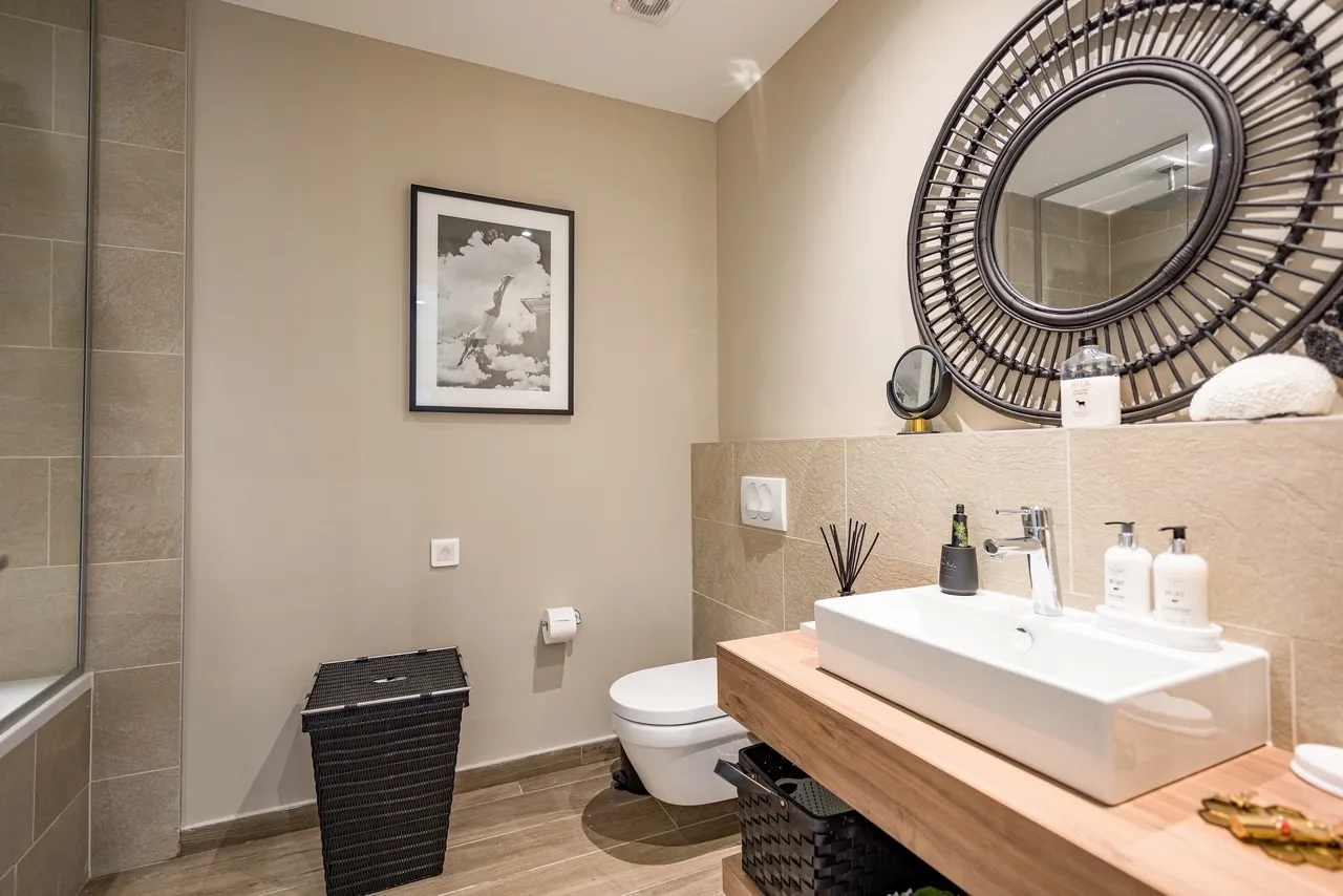 Modern bathroom featuring a wall-mounted toilet, rectangular vessel sink on a wooden vanity, and a large round decorative mirror.