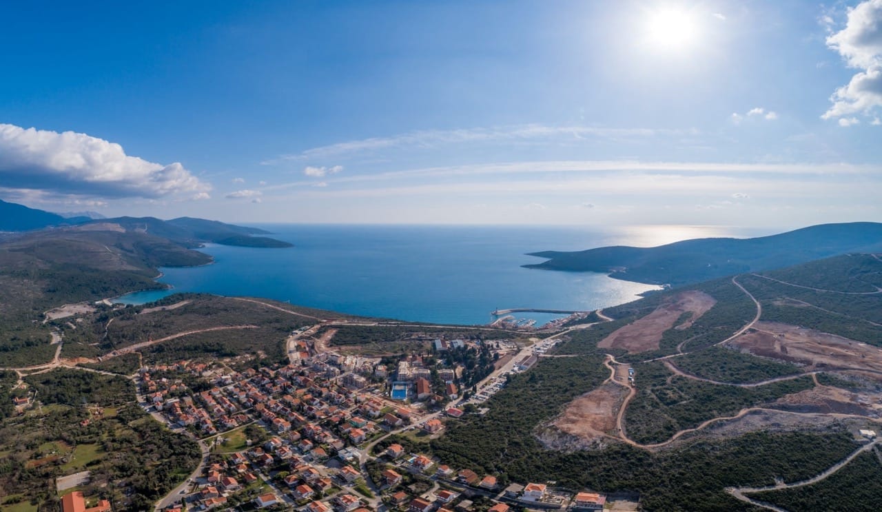 Aerial view of a coastal town with red roofs, surrounded by green hills and blue sea under bright sun.