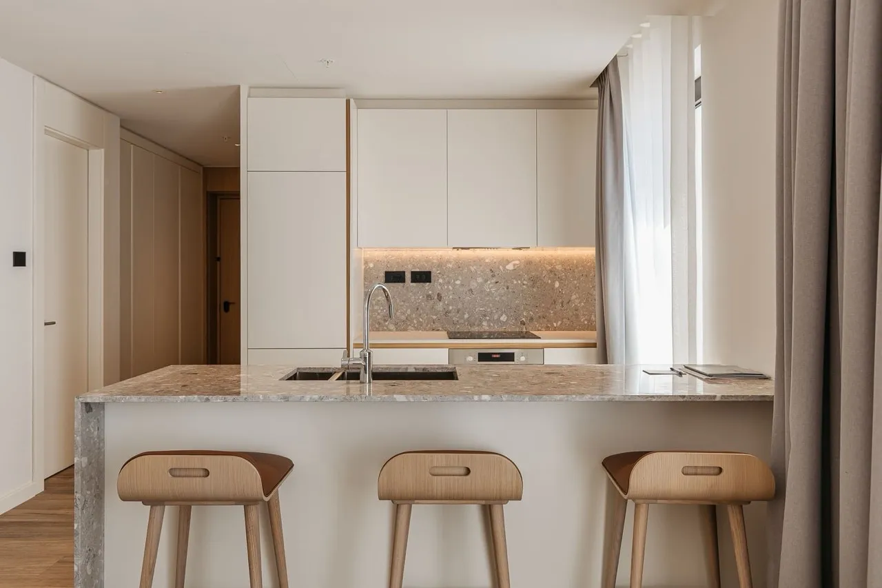 Modern kitchen featuring a stone island with sink, wooden bar stools, and white cabinetry.