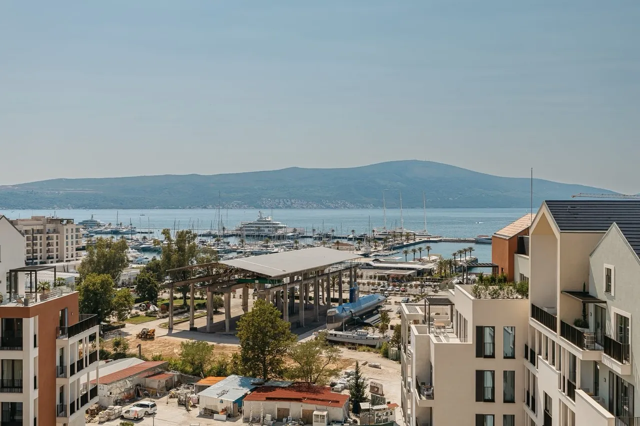 View of a yacht marina with boats and mountains in the background. Modern buildings and a construction area are visible.