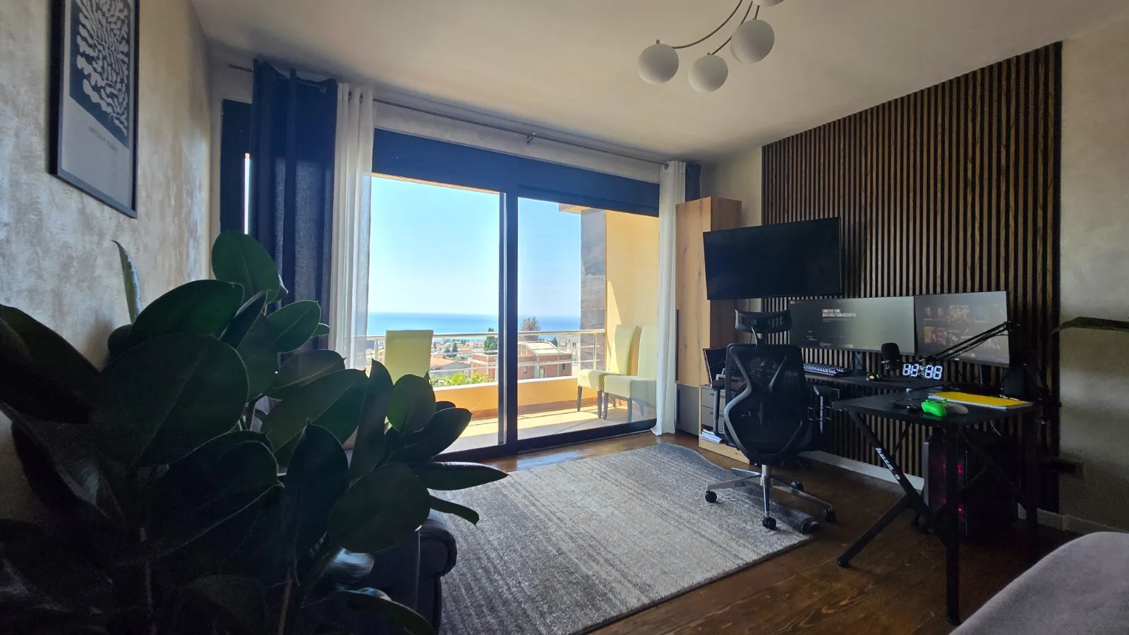 Room featuring a computer desk, multiple monitors, and gaming chair near a sliding glass door with a sea view.