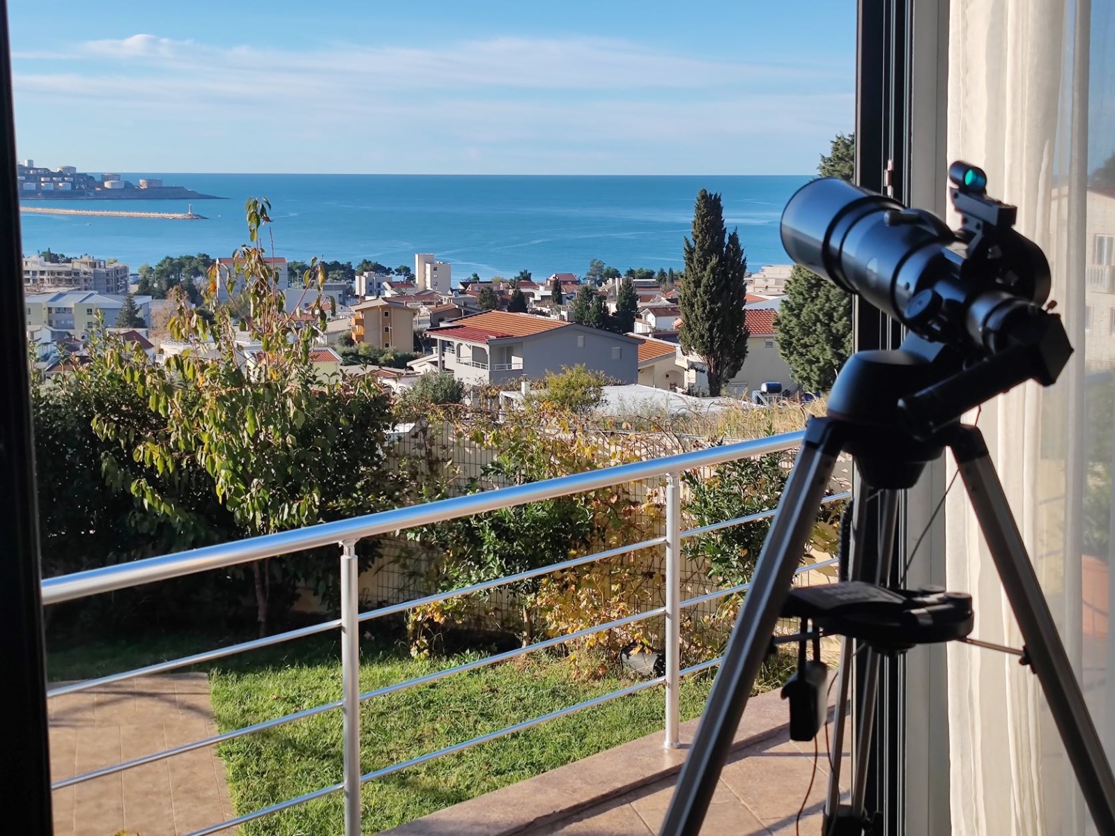 View from the villa balcony in Bar, Montenegro: a panorama of the sea and the city, a telescope on a tripod, a terrace with a railing.