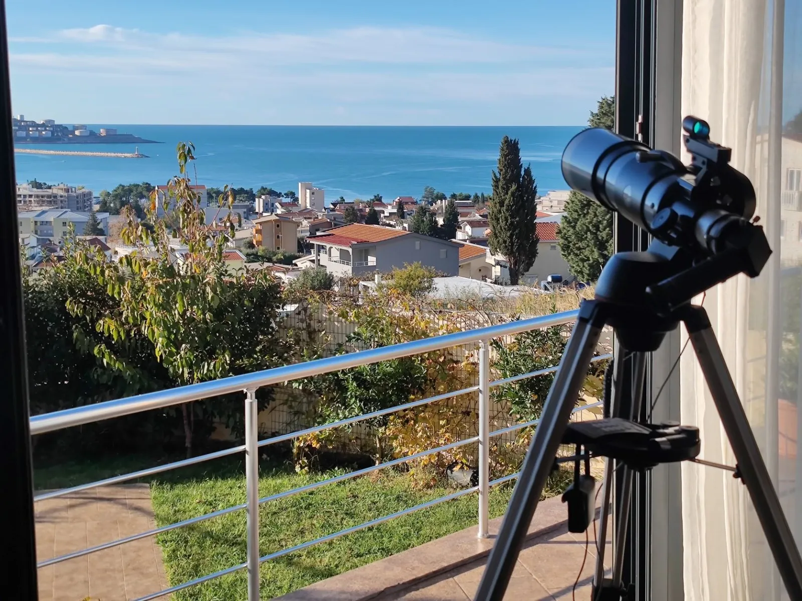 Telescope on a balcony overlooking the sea and city. Metal railing and green area visible.