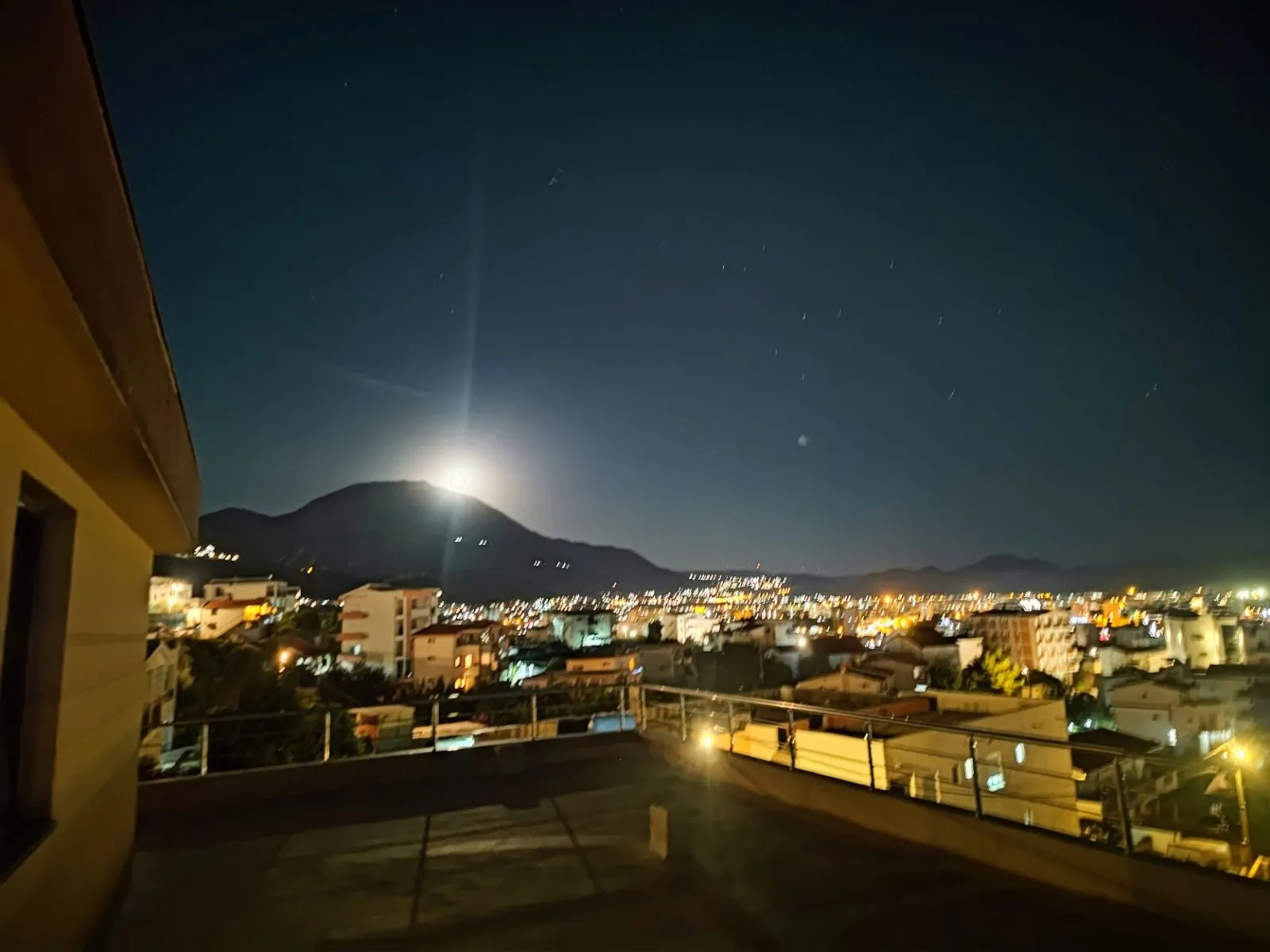 Night view from a terrace showing city lights, a mountain silhouette under the moon, and a railing.