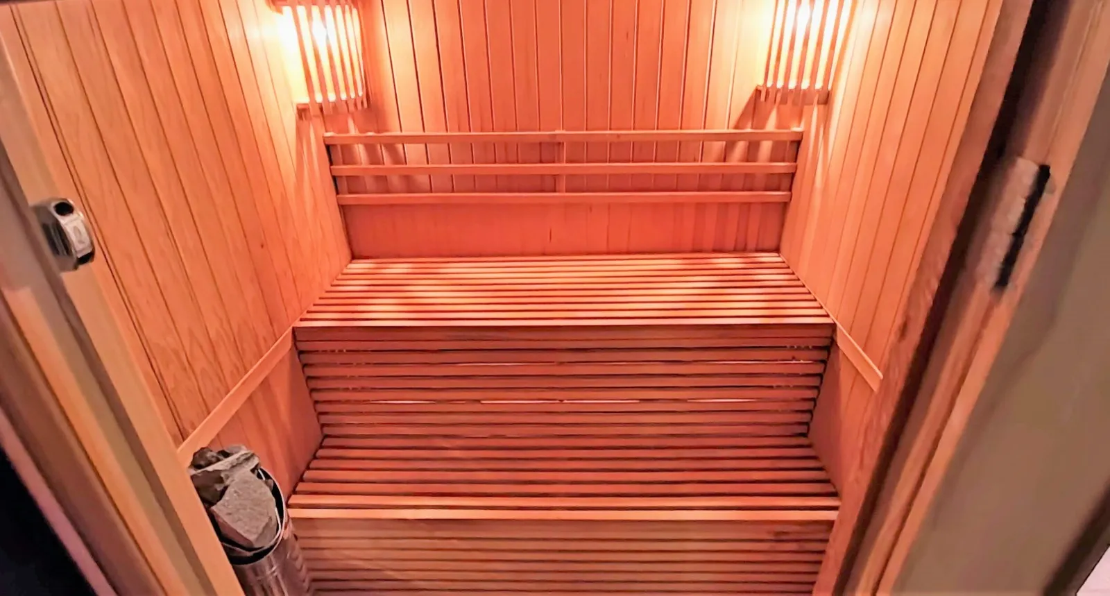 Wooden sauna with tiered benches, heaters on walls, and a bucket containing sauna stones.