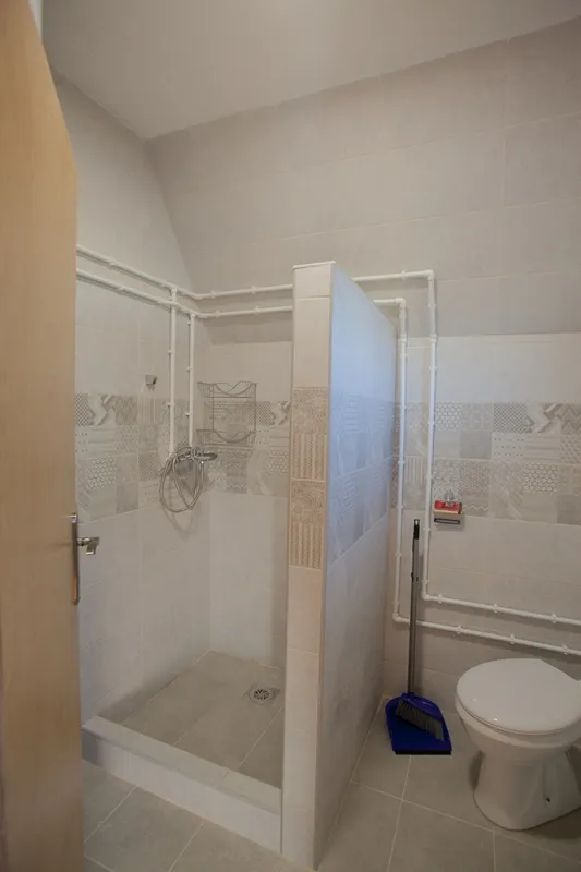 Bathroom with a shower stall, toilet, and exposed white pipes on tiled walls.