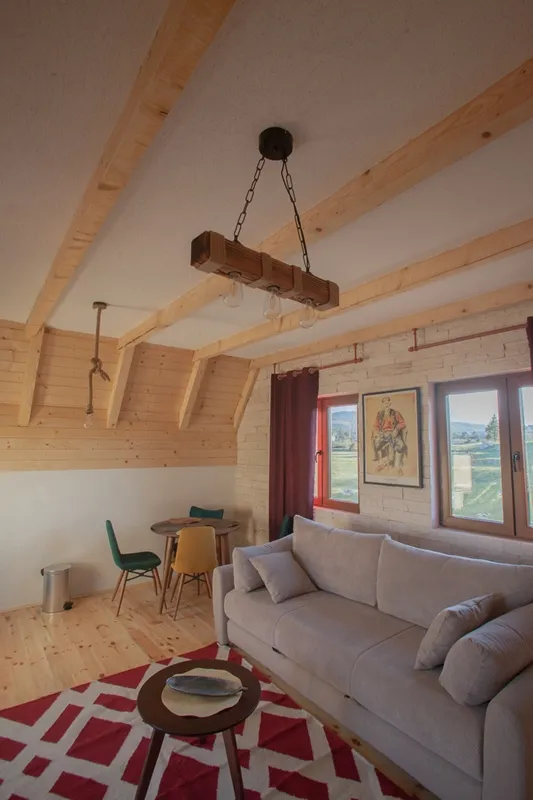 Living room featuring a grey sofa, wooden beam ceiling, and a small dining area near windows.