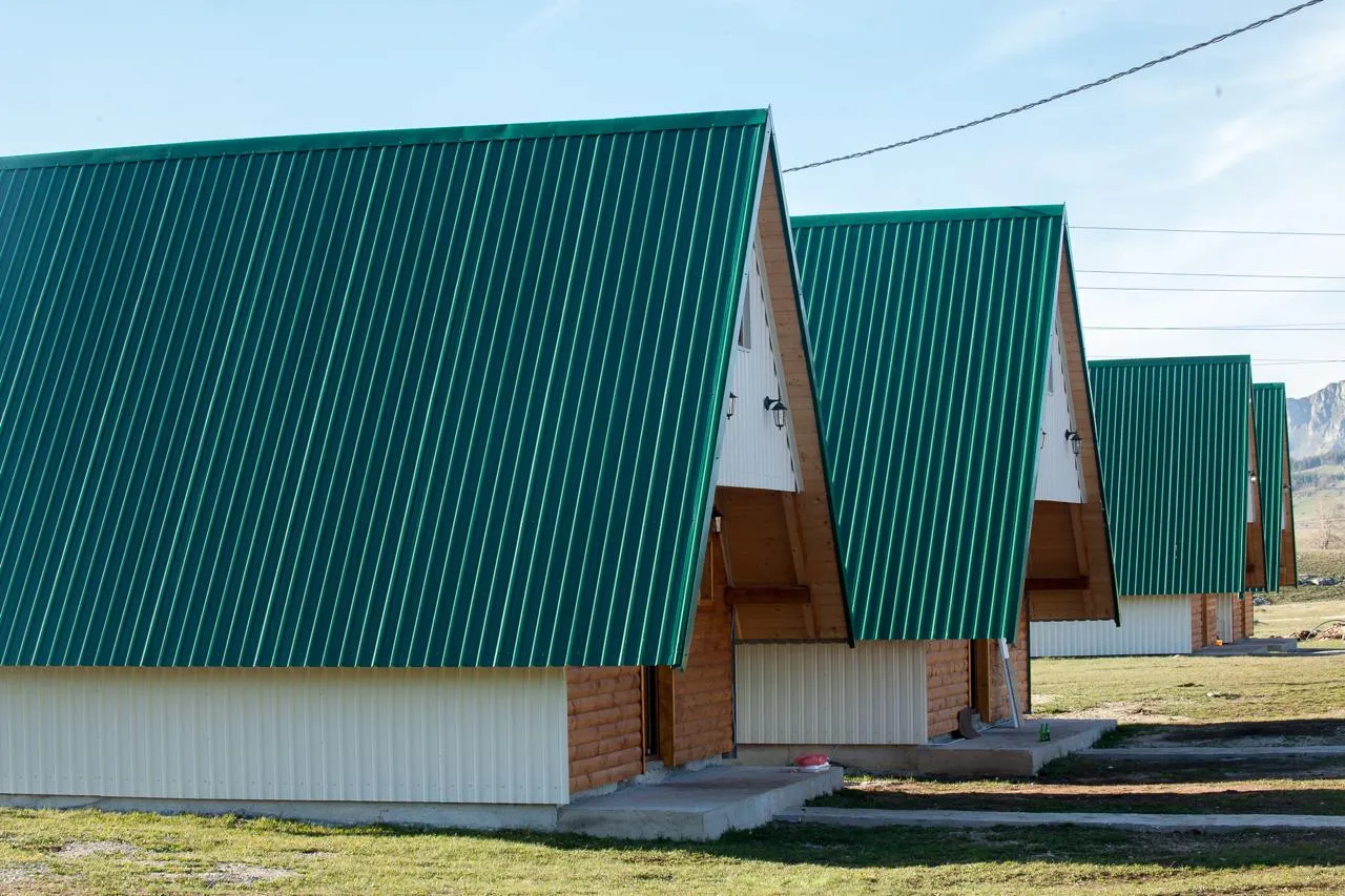 Row of wooden cabins with green roofs set against grass and mountains.