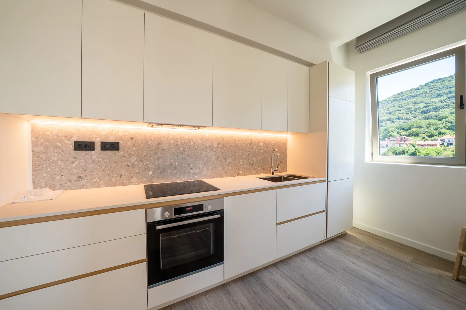 Modern kitchen with white cabinets, terrazzo backsplash, built-in oven, sink, and a window overlooking hills.
