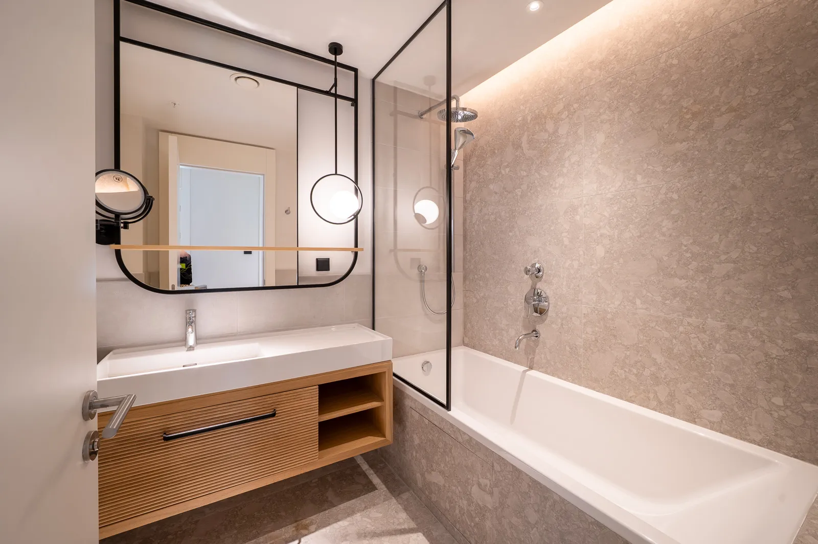 Modern bathroom featuring a wooden vanity, white sink, large mirror, and bathtub with shower on terrazzo tiles.